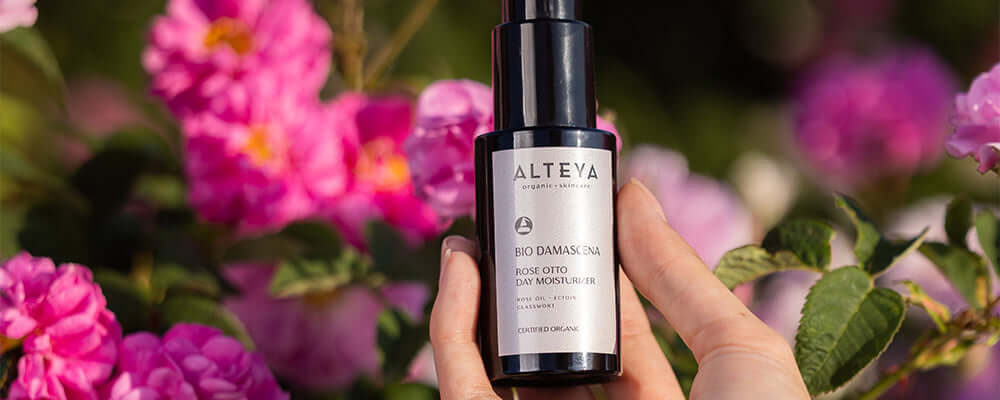 A hand holding a bottle of facial oil in front of flowers.