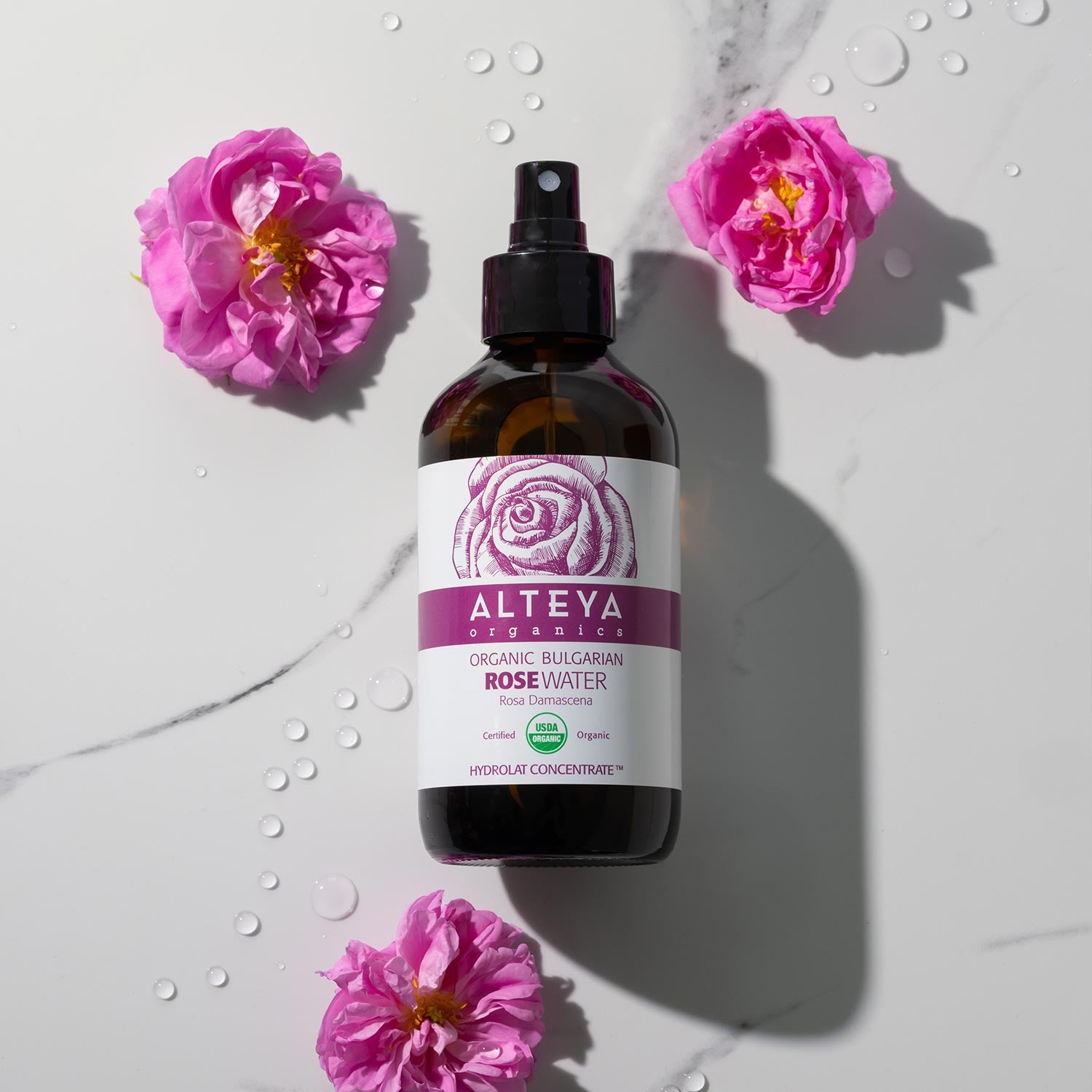 A bottle of Bulgarian Organic Rose Water – Glass Spray by Alteya Organics, known for its hydrating properties, surrounded by a bed of pink flowers.