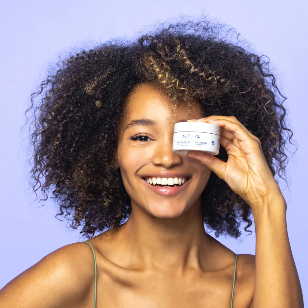 A woman with curly hair holding a jar of Rose Hydrobiome Bakuchiol Barrier Cream.
