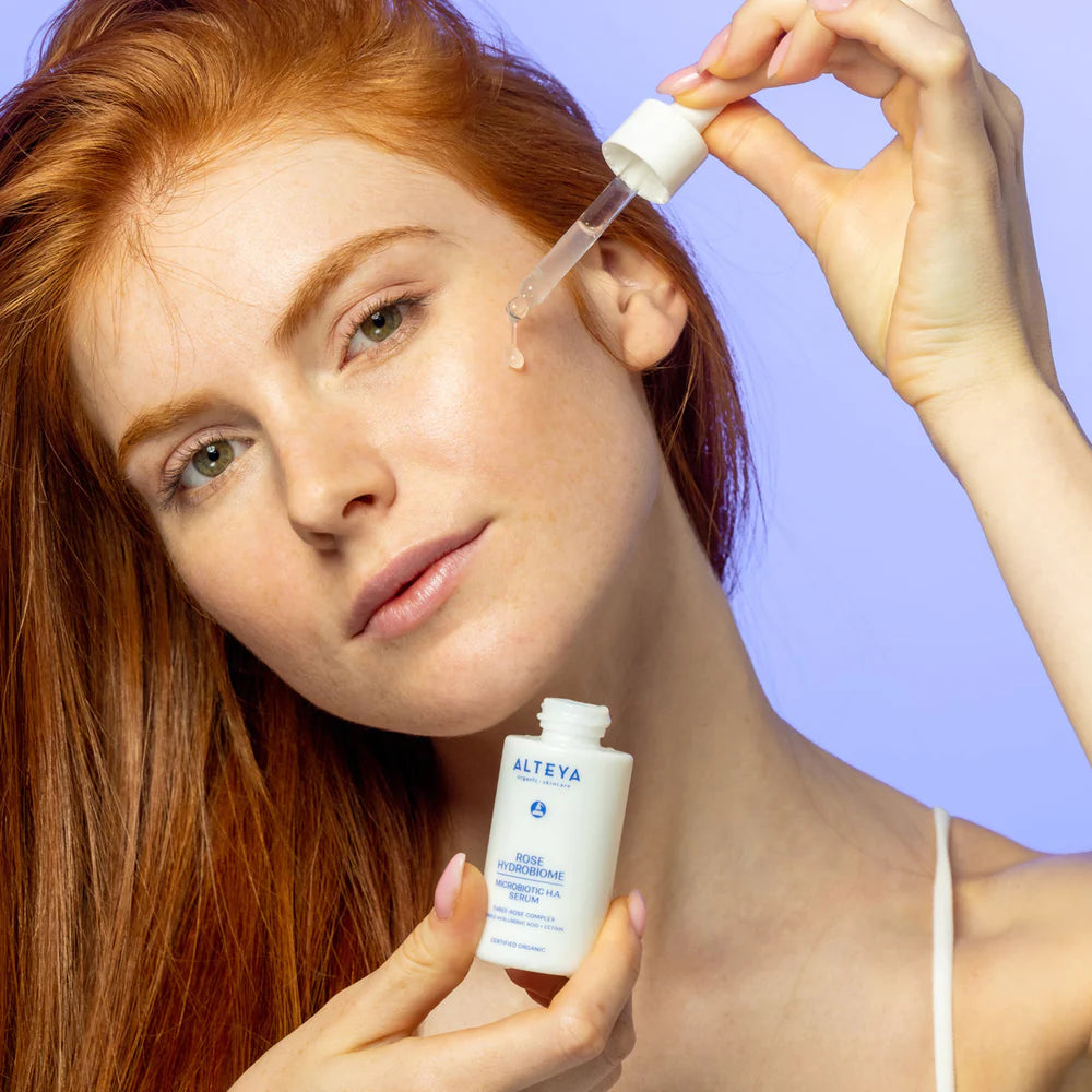 A woman with red hair is holding a bottle of Rose Hydrobiome Microbiotic H.A. Serum.