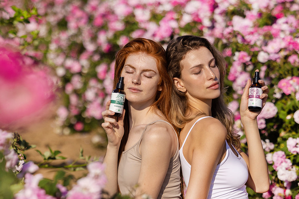Two women holding bottles of essential oils in a field of roses.