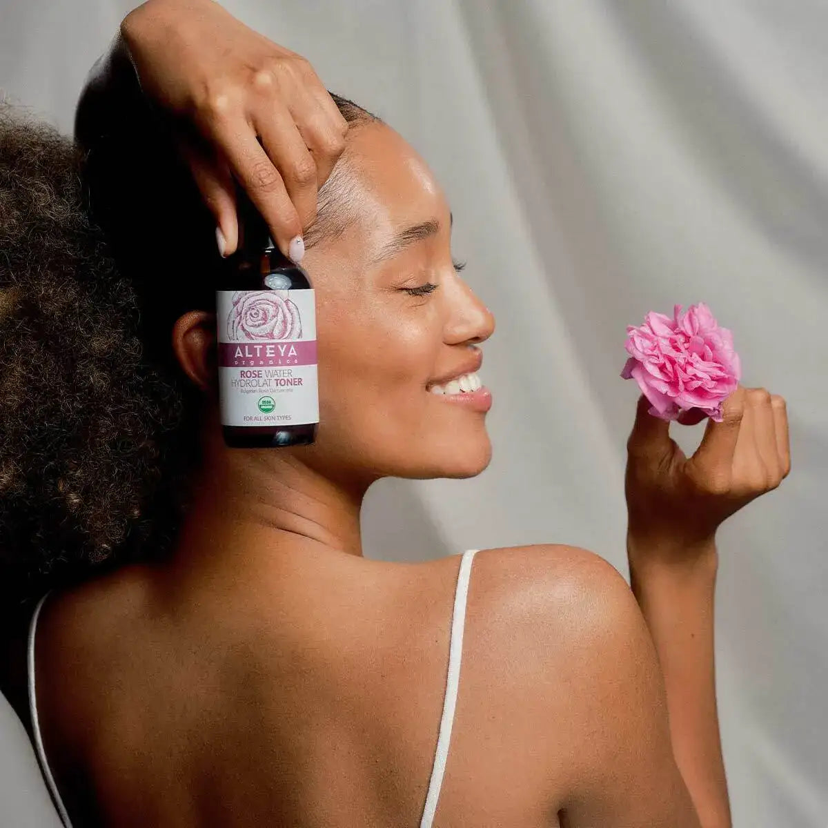 A woman holds a bottle labeled "Bulgarian Organic Rose Water – Glass Spray" near her head and a pink rose in her other hand, smiling with eyes closed against a neutral background. Made with organic rose water from the Bulgarian Rose Valley, this product is designed to hydrate and refresh the skin.