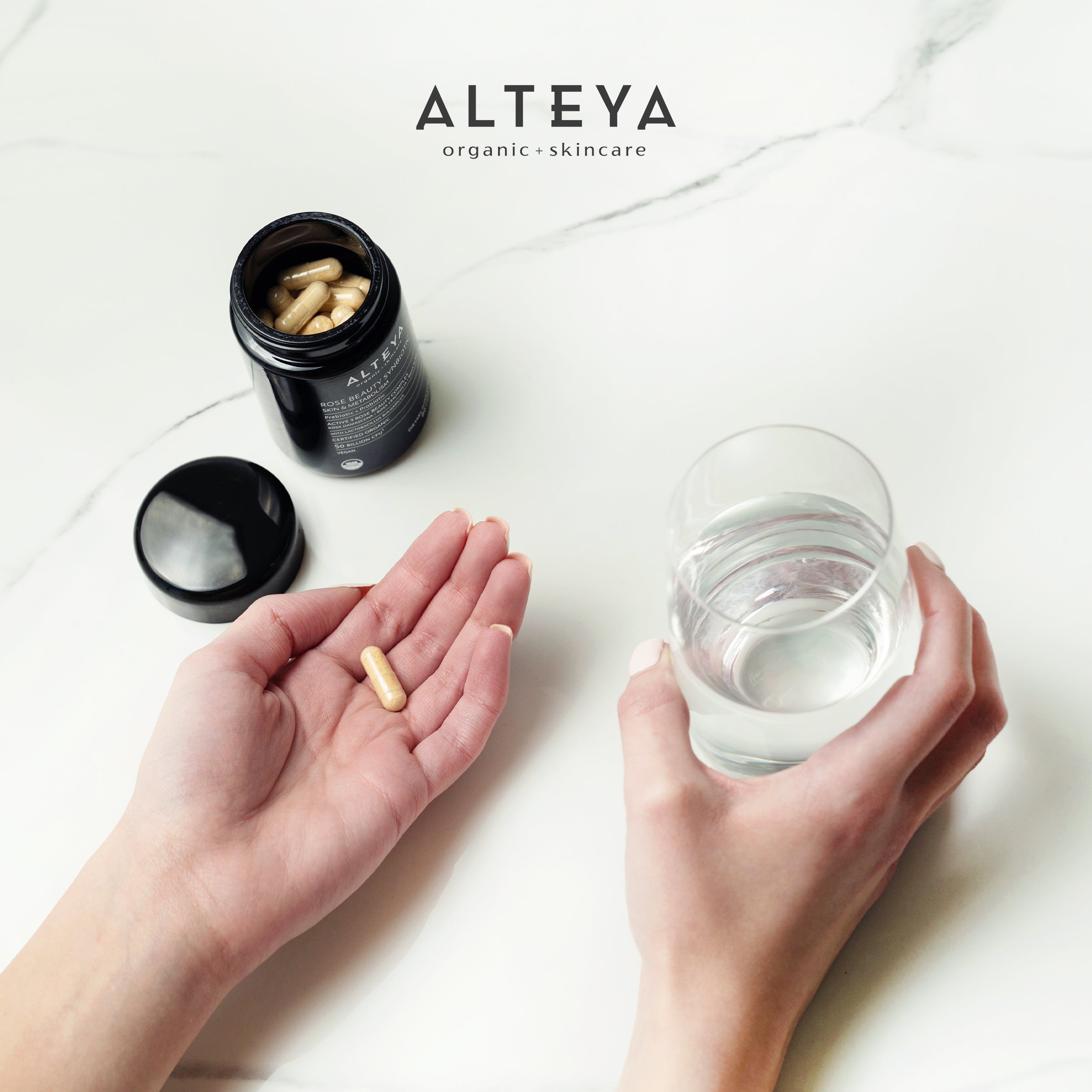 A woman's hand holding a glass of water and a bottle of Rose Beauty Prebiotic and Probiotic - Synbiotic Skin & Metabolism Vegan Organic Supplement by Alteya Organics for gut health.