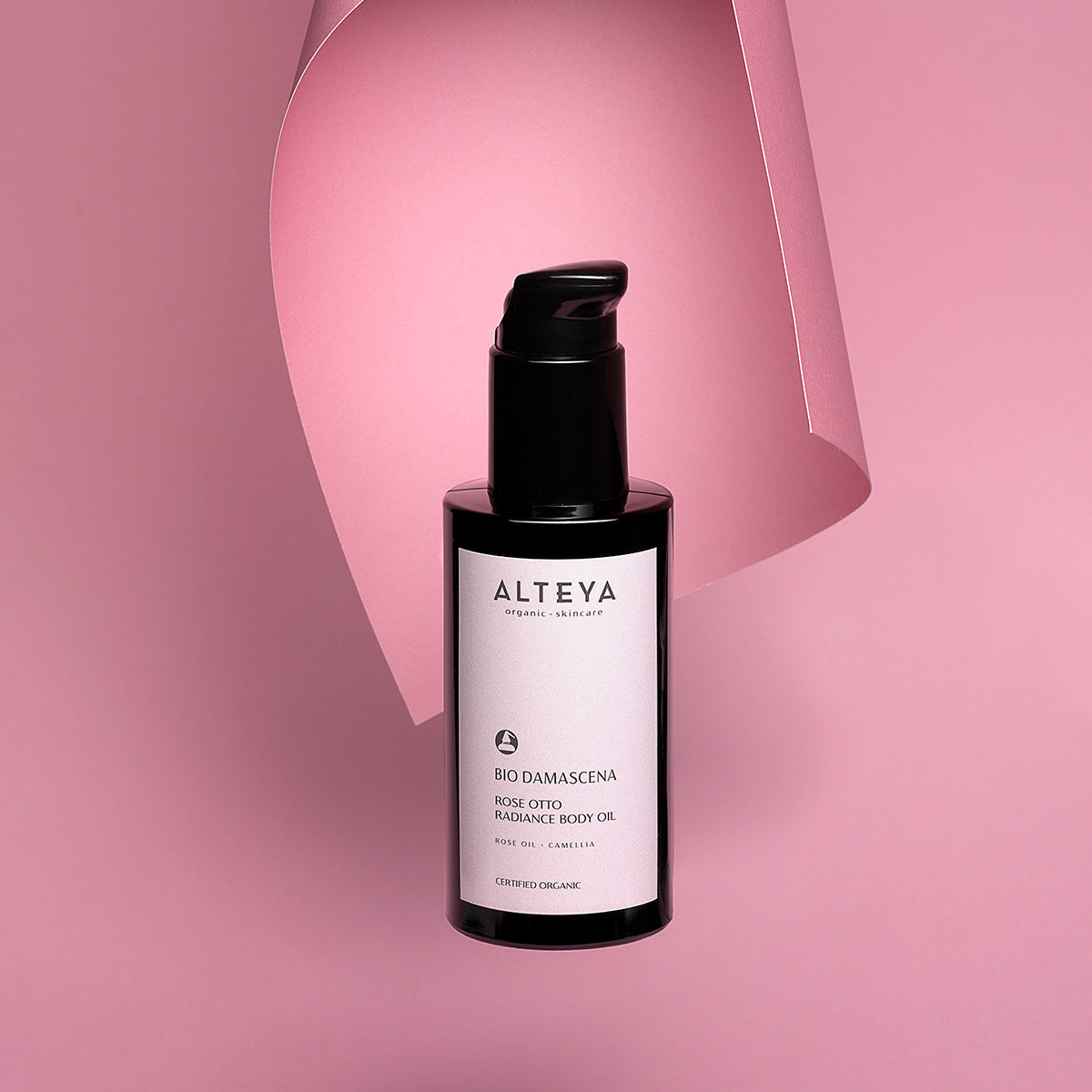 A bottle of Alteya Organics Bio Damascena Rose Otto Radiance Body Oil, rich in essential nutrients, on a pink background.