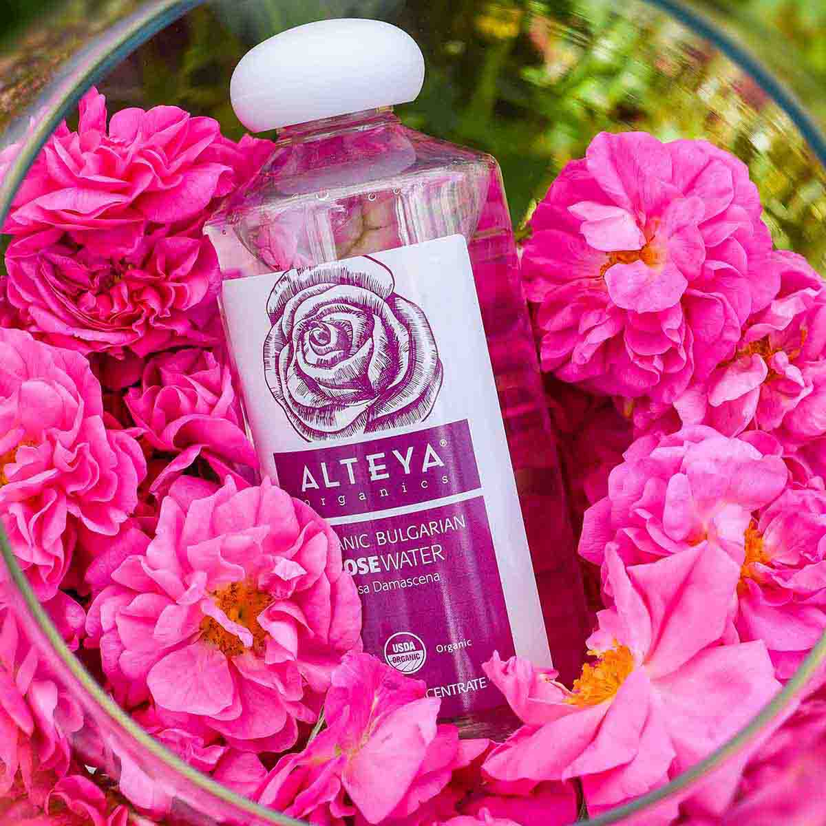 A bottle of Alteya Organics Organic Bulgarian Rose Water - 17 Fl Oz, perfect for skincare hydration, surrounded by pink flowers.