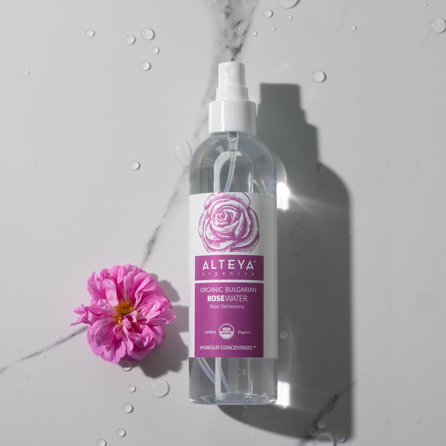 A bottle of Alteya Organics Organic Bulgarian Rose Water Toner Mist - 8.5 Fl Oz next to a pink flower.