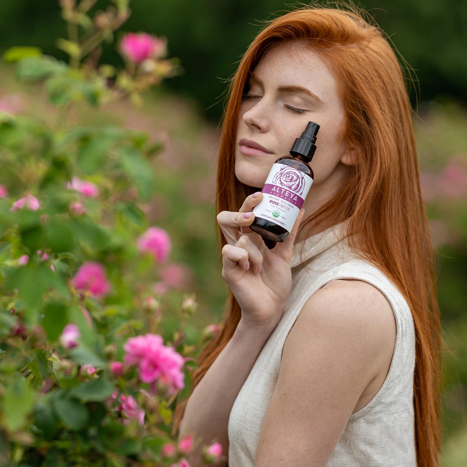 A woman with red hair is holding a bottle of Alteya Organics' Bulgarian Organic Rose Water – Glass Spray, providing hydration for her skincare routine.