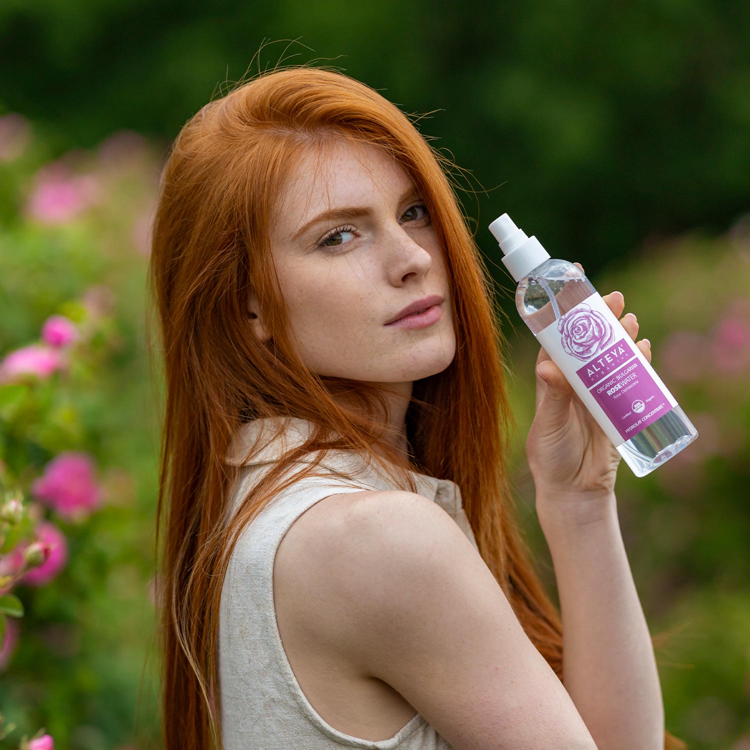 A woman with vibrant red hair holding a bottle of Alteya Organics' Organic Bulgarian Rose Water Toner Mist - 8.5 Fl Oz spray.